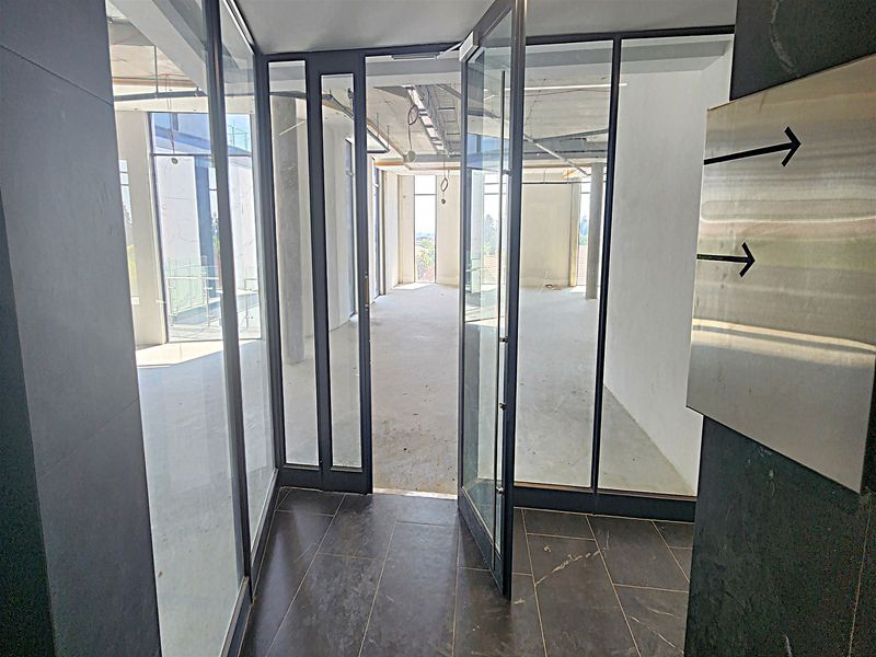 An unfinished open-plan office space featuring glass partitions, exposed ceiling infrastructure, and dark floor tiling, currently in a shell condition.