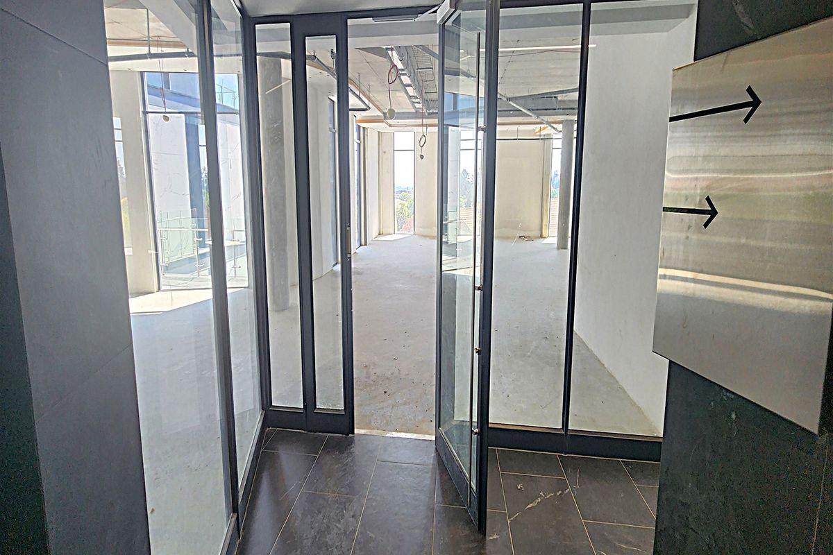 An unfinished open-plan office space featuring glass partitions, exposed ceiling infrastructure, and dark floor tiling, currently in a shell condition.