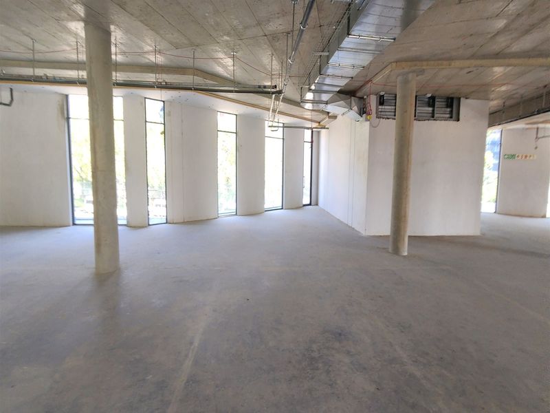 An expansive, open-plan office space in shell condition, featuring exposed concrete ceilings, support columns, and large windows providing ample natural light.