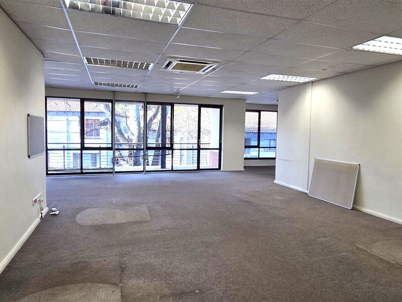 A spacious, empty open-plan office area featuring suspended ceilings, recessed lighting, and large windows providing ample natural light.