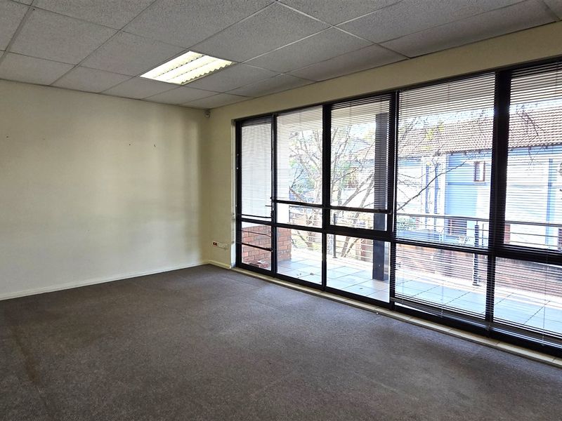 An empty office space featuring a suspended ceiling, recessed lighting, and large windows with fitted blinds, offering ample natural light and a balcony view.