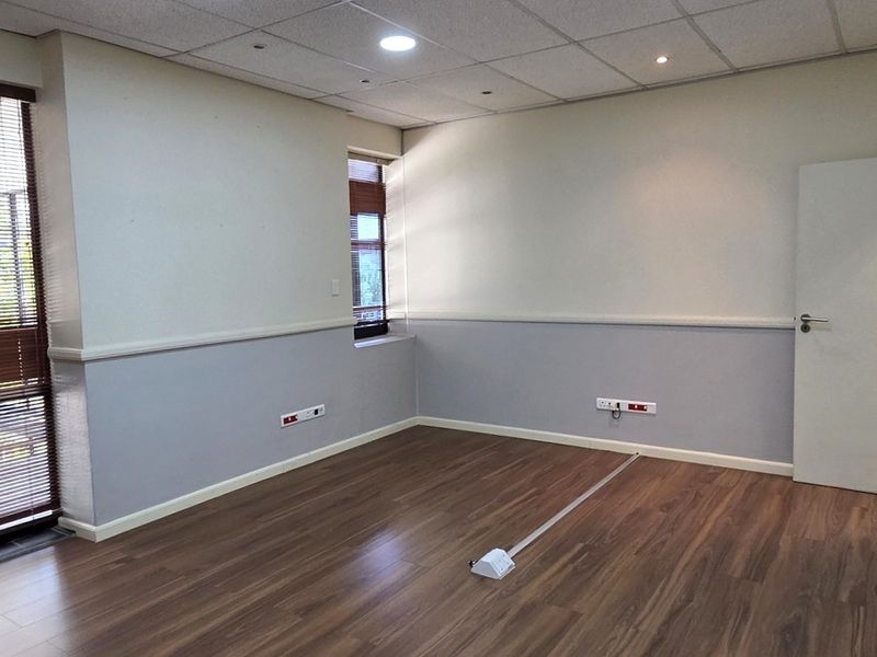 A clean, empty office space featuring laminate flooring, a suspended ceiling with recessed lighting, and large windows with fitted venetian blinds.