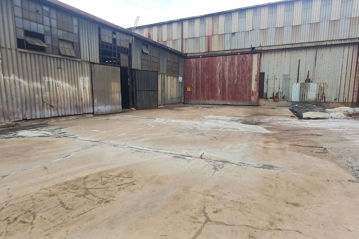 Industrial yard area featuring corrugated metal warehouse structures with large sliding access doors and significant signs of weathering and surface wear.