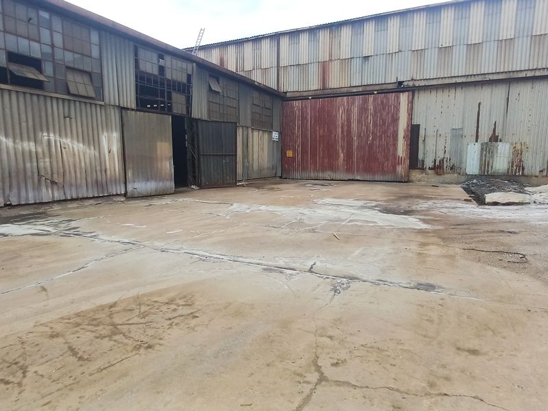Industrial yard area featuring corrugated metal warehouse structures with large sliding access doors and significant signs of weathering and surface wear.
