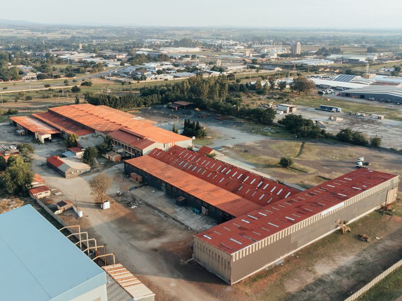 Aerial view of an industrial facility featuring multiple large warehouses with metal roofing, expansive yard space, and perimeter fencing in an industrial zone.