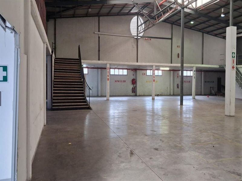 Spacious industrial warehouse area featuring concrete flooring, a mezzanine level with a steel staircase, and exposed roof trusses, suitable for storage or manufacturing operations.
