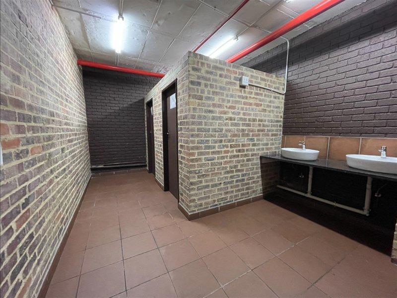 Industrial-style communal bathroom featuring exposed brick walls, tiled flooring, dual vanity basins, and exposed fire sprinkler piping.
