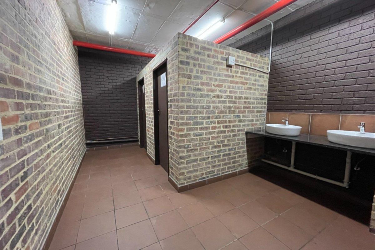 Industrial-style communal bathroom featuring exposed brick walls, tiled flooring, dual vanity basins, and exposed fire sprinkler piping.