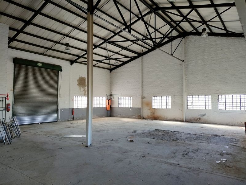 Spacious industrial warehouse unit featuring high ceilings with exposed steel trusses, concrete flooring, and a large roller shutter door for easy access.