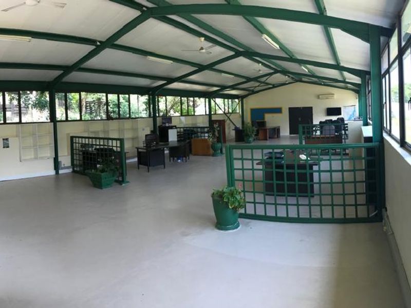 Spacious open-plan office area featuring high ceilings with exposed steel trusses, extensive natural light through large windows, and functional metal security partitions.