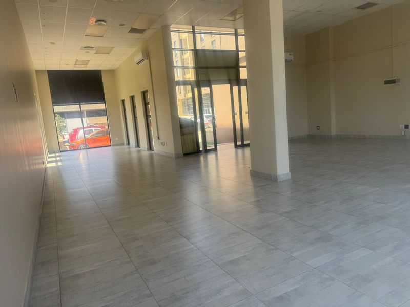 Spacious open-plan retail unit featuring large glass storefront windows, tiled flooring, suspended ceiling, and wall-mounted air conditioning units.