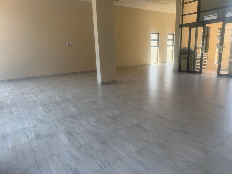 A spacious, open-plan retail unit featuring modern wood-look tiled flooring, neutral walls, and large glass entrance doors providing ample natural light.