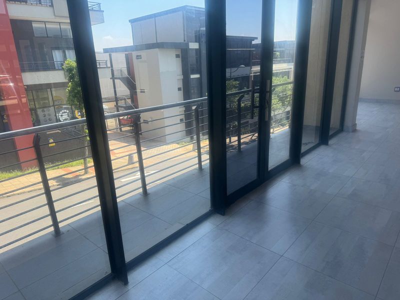 A bright interior space featuring large glass sliding doors that open onto a balcony with metal railings, overlooking the street and surrounding commercial buildings.