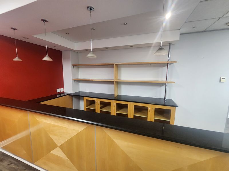 A clean, empty office bar area featuring a built-in wooden counter with a granite top, open shelving, and modern pendant lighting.