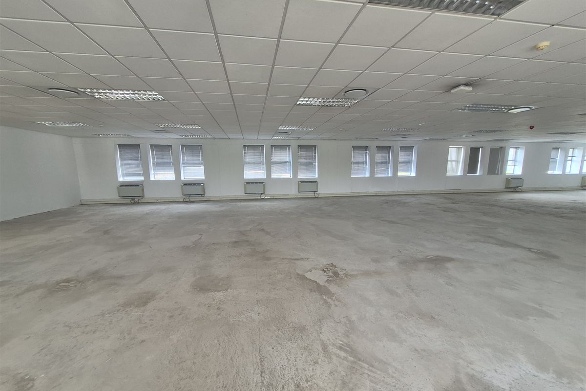 A large, open-plan office space featuring a suspended ceiling, recessed lighting, wall-mounted air conditioning units, and unfinished concrete flooring.