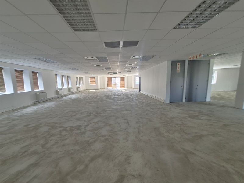 A spacious, open-plan office area featuring a suspended ceiling, recessed lighting, air conditioning units, and large windows with venetian blinds.
