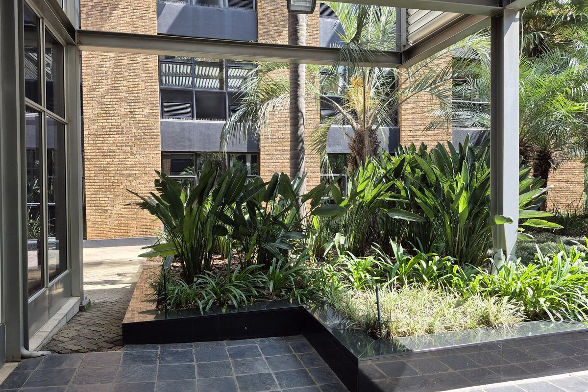 A covered outdoor patio area featuring stone tiling, a modern steel pergola structure, and a raised garden planter against a brick office building facade.