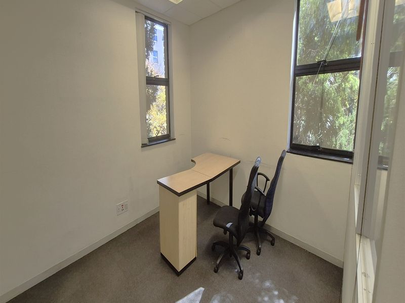 A compact office space featuring carpeted flooring, suspended ceiling tiles, and large windows providing natural light.