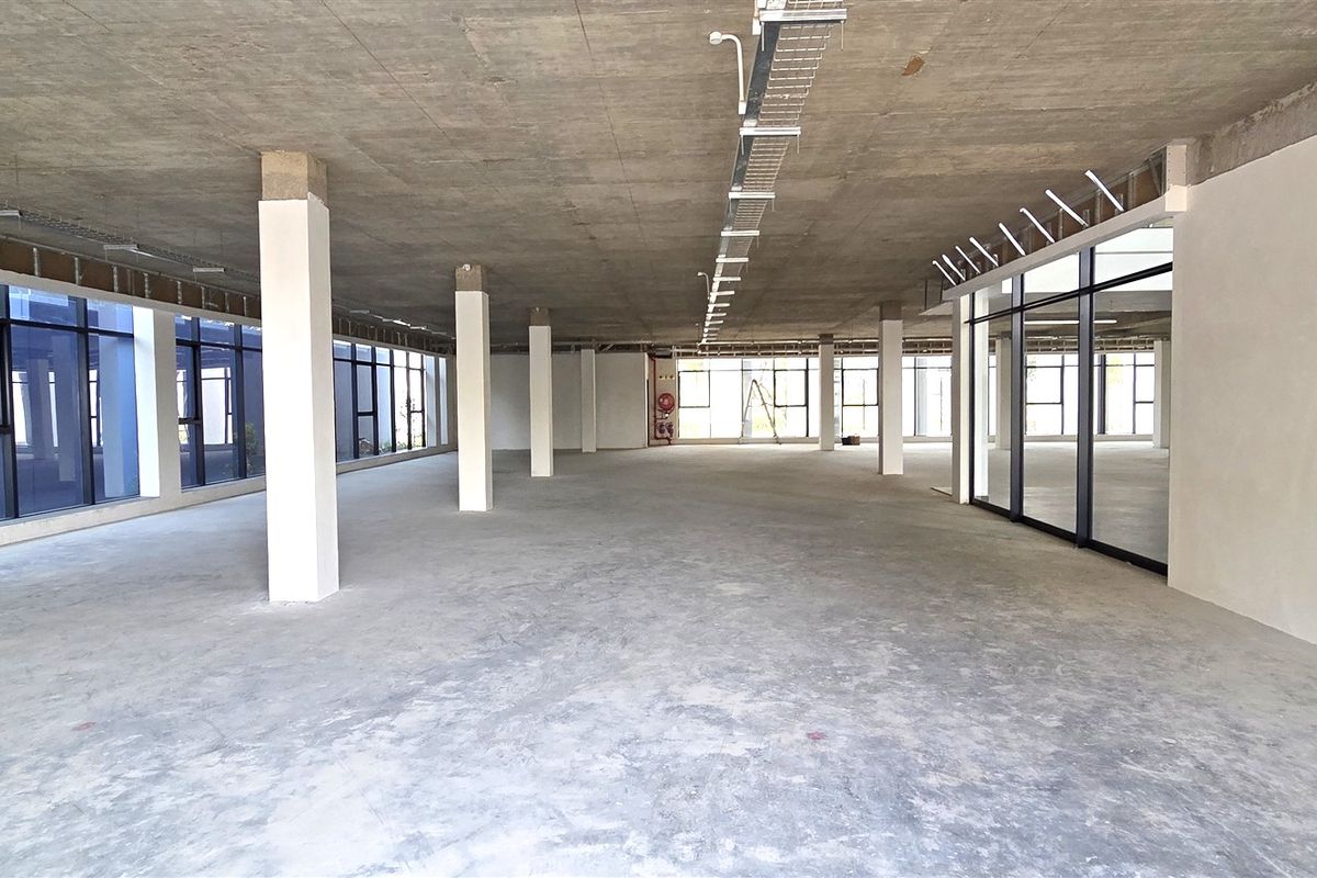 A large, open-plan office space in shell condition featuring exposed concrete ceilings, structural columns, and extensive floor-to-ceiling windows providing ample natural light.