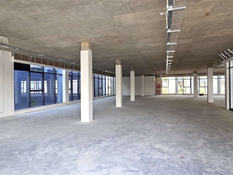 A spacious, open-plan office area featuring an exposed concrete ceiling, large windows providing natural light, and structural support columns in a shell condition.