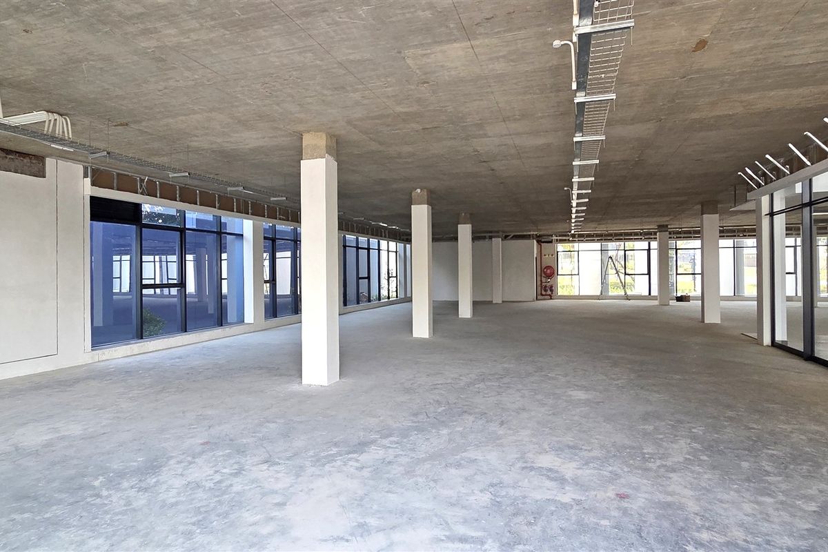 A spacious, open-plan office area featuring an exposed concrete ceiling, large windows providing natural light, and structural support columns in a shell condition.