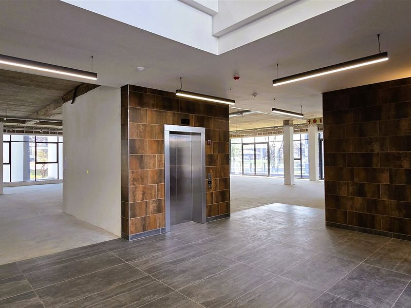 Spacious open-plan office area featuring modern tiled flooring, an elevator, and large windows providing ample natural light with an industrial-style exposed concrete ceiling.