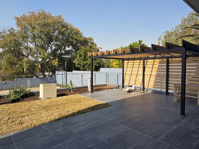 A modern outdoor patio area featuring a timber pergola, glass balustrade, and dark tiled flooring, overlooking a landscaped garden space.