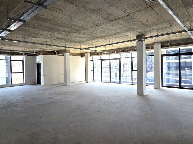 Spacious open-plan office area featuring exposed concrete ceilings, structural columns, and large floor-to-ceiling windows providing ample natural light.