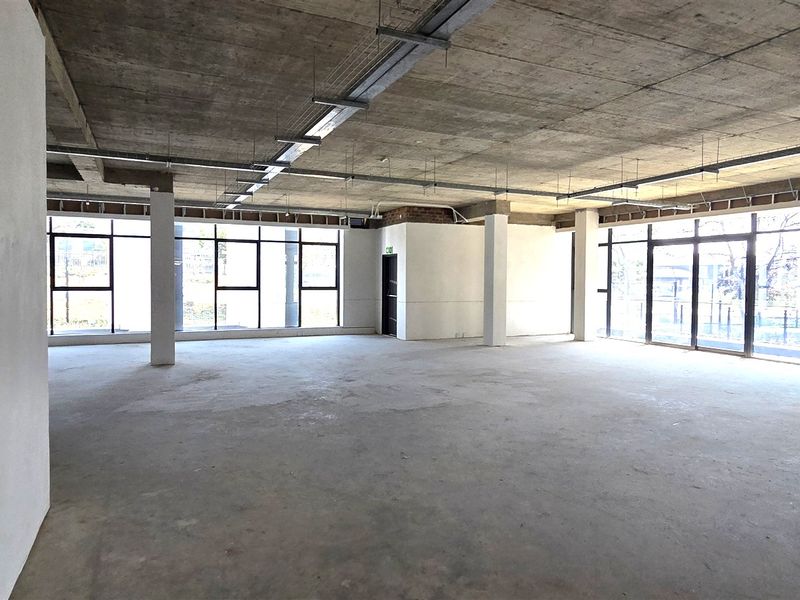 A spacious, open-plan office area featuring an industrial aesthetic with exposed concrete ceilings, structural columns, and large windows providing ample natural light.