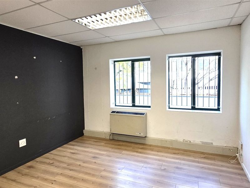 An empty office space featuring laminate flooring, a suspended ceiling with recessed lighting, security-barred windows, and a wall-mounted HVAC unit.