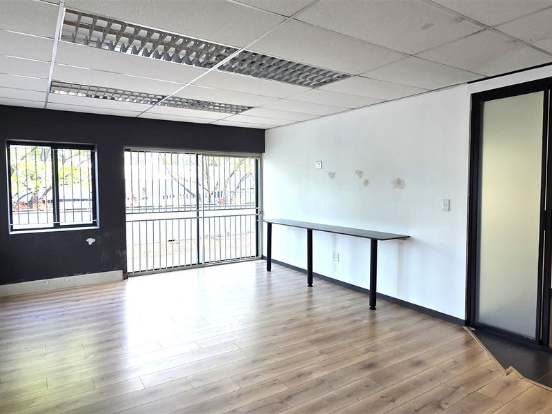 A bright, empty office space featuring laminate flooring, a built-in desk, security bars on windows, and a modern glass partition leading to an adjacent area.