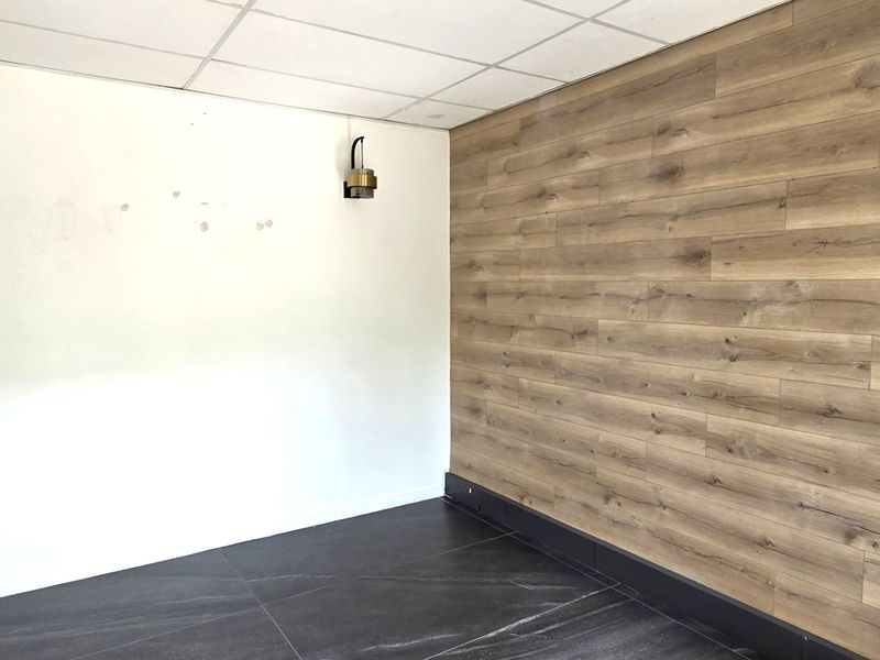 A vacant office space featuring a suspended ceiling, wood-look accent wall, dark tiled flooring, and modern wall-mounted lighting fixtures.