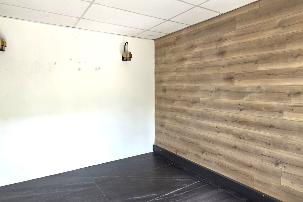 A vacant office space featuring a suspended ceiling, wood-look accent wall, dark tiled flooring, and modern wall-mounted lighting fixtures.