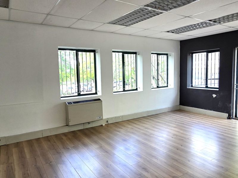 A bright, empty office space featuring laminate flooring, a suspended ceiling with recessed lighting, and large windows with security bars.