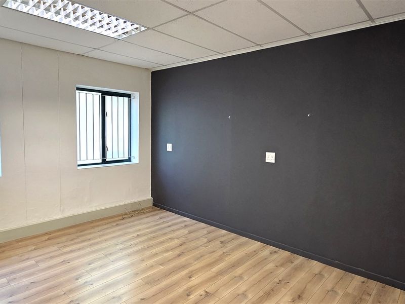 A vacant office space featuring laminate flooring, a suspended ceiling with recessed lighting, security bars on windows, and a wall-mounted air conditioning unit.