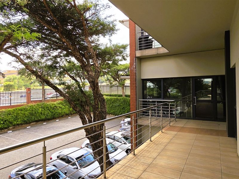 A spacious covered balcony featuring tiled flooring and a modern stainless steel balustrade, overlooking the office parking area and landscaped grounds.