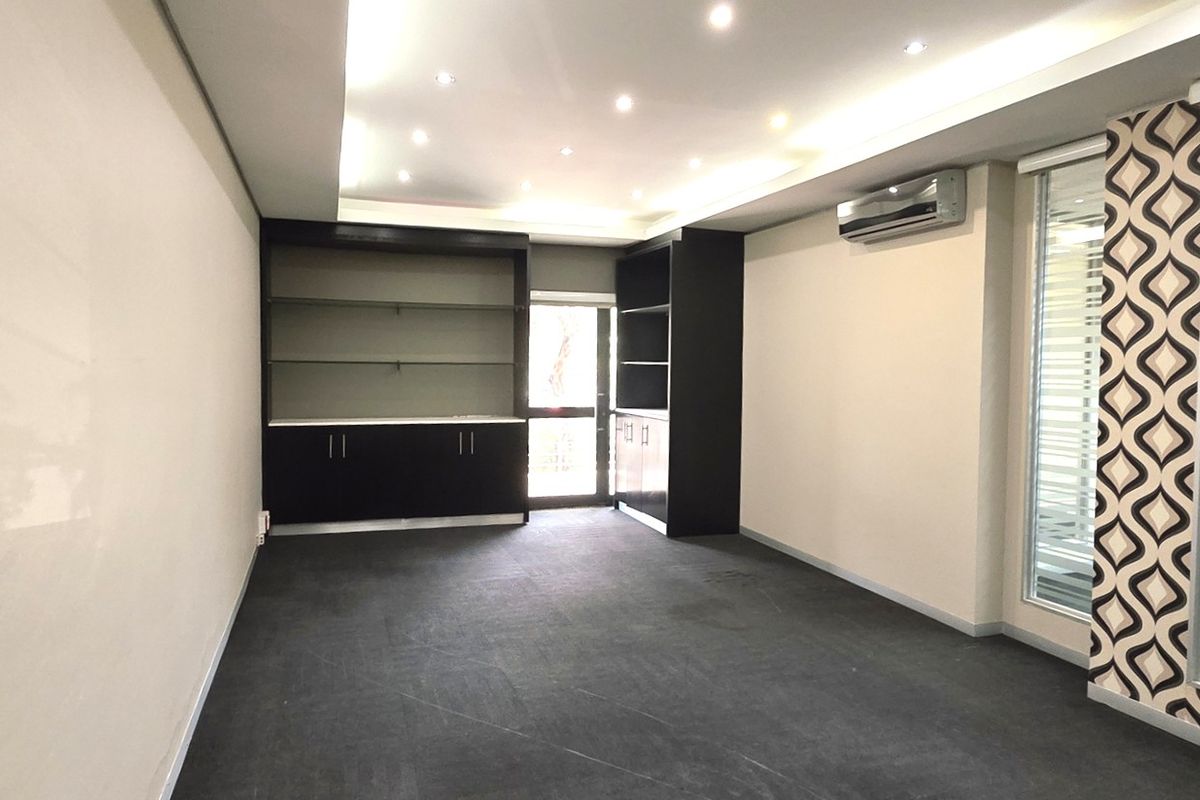 A clean, empty office space featuring built-in storage cabinetry, recessed ceiling lighting, a wall-mounted air conditioning unit, and modern glass partitions.