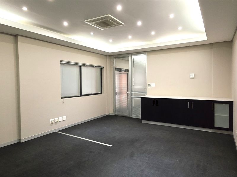 A clean, empty office space featuring recessed ceiling lighting, ducted air conditioning, dark carpet flooring, and built-in storage cabinetry.