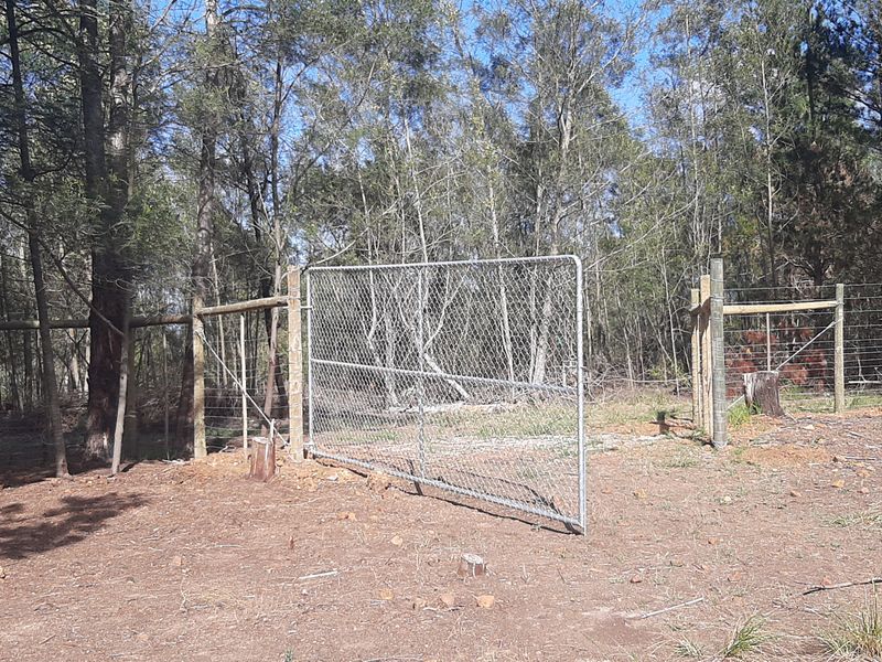 A rural farm property featuring a chain-link gate and wire fencing set against a backdrop of established trees and natural woodland.