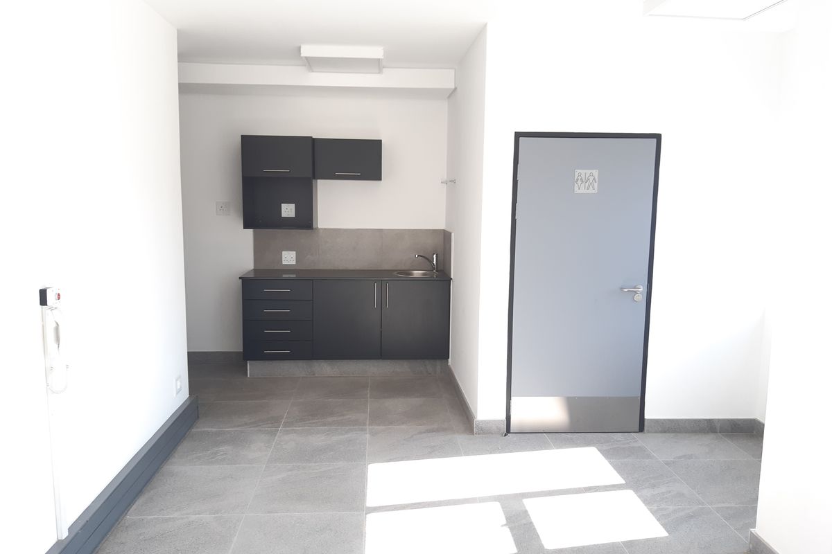 A clean, modern kitchenette area featuring dark cabinetry, a stainless steel sink, and neutral tiled flooring, adjacent to a private restroom door.