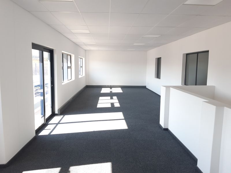 A bright, open-plan office space featuring neutral finishes, carpeted flooring, a suspended ceiling, and large windows providing ample natural light.