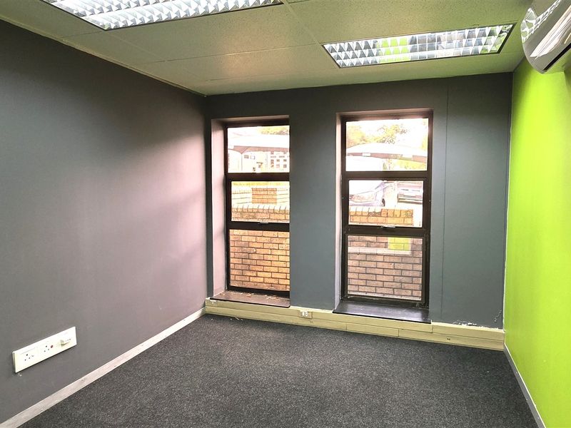 A vacant office space featuring carpeted flooring, a suspended ceiling with recessed lighting, a wall-mounted air conditioner, and dual windows with aluminium frames.