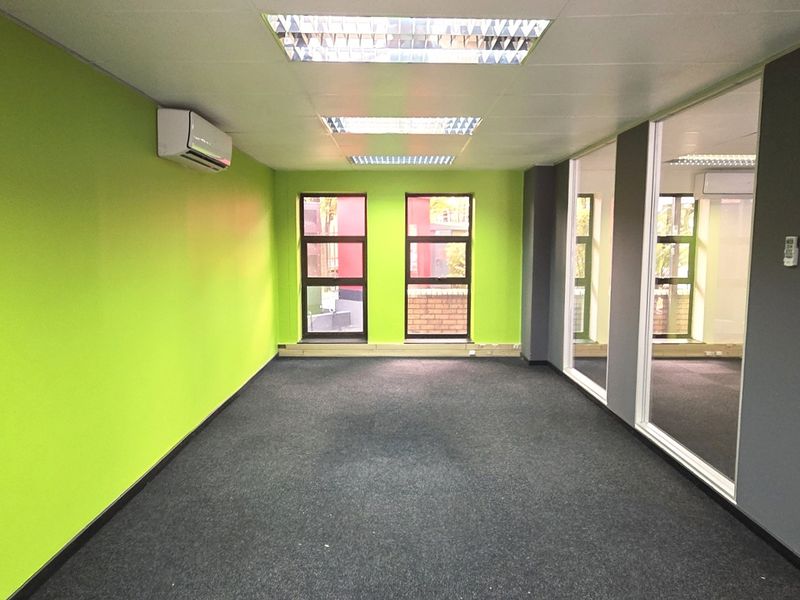 A bright, empty office space featuring green accent walls, glass partitions, carpeted flooring, and integrated air conditioning.