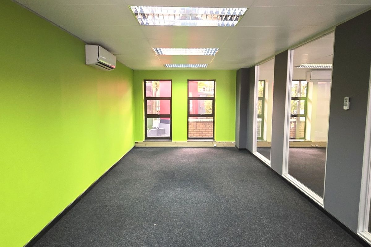 A bright, empty office space featuring green accent walls, glass partitions, carpeted flooring, and integrated air conditioning.