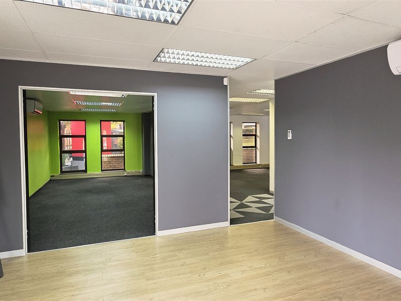 A bright, open-plan office space featuring neutral grey walls, a mix of laminate and carpet flooring, and integrated air conditioning.