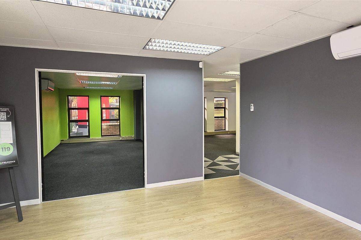 A bright, open-plan office space featuring neutral grey walls, a mix of laminate and carpet flooring, and integrated air conditioning.