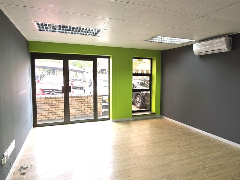 A bright, empty office space featuring laminate flooring, a suspended ceiling with recessed lighting, and a wall-mounted air conditioning unit.