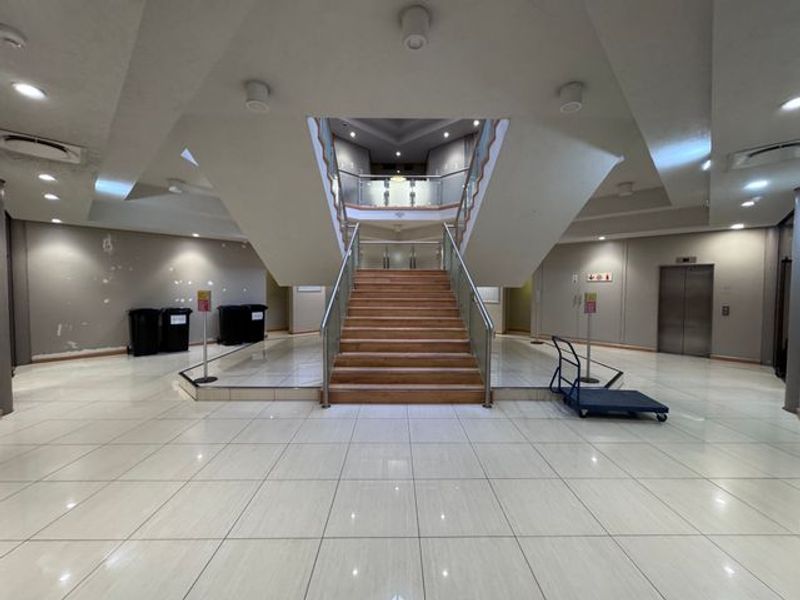 Spacious industrial building entrance lobby featuring a central staircase with glass balustrades, tiled flooring, and elevator access.