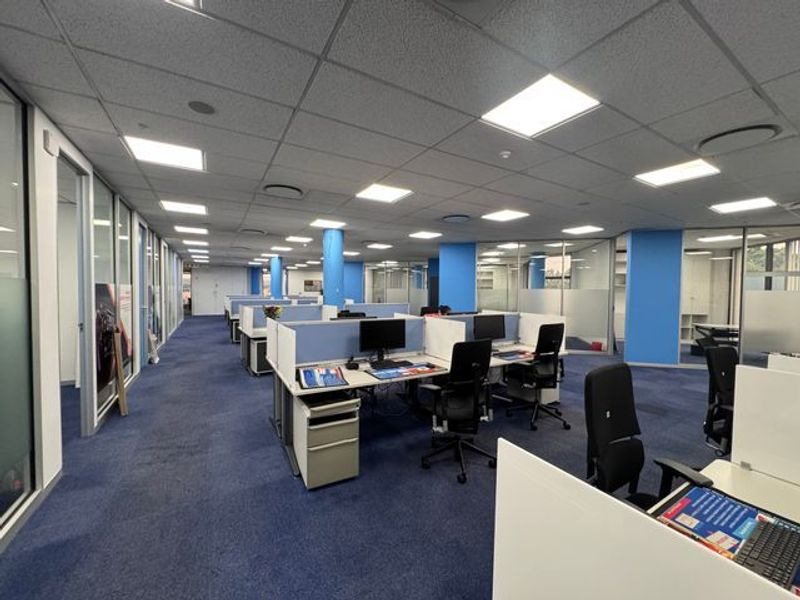 Open-plan office space featuring glass partitions, suspended ceilings with recessed lighting, and blue carpet flooring, providing a professional and functional work environment.