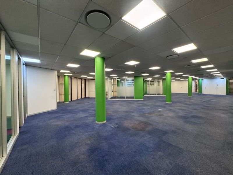Expansive open-plan industrial office space featuring a suspended ceiling, recessed lighting, structural columns, and glass-partitioned offices with blue carpet flooring.